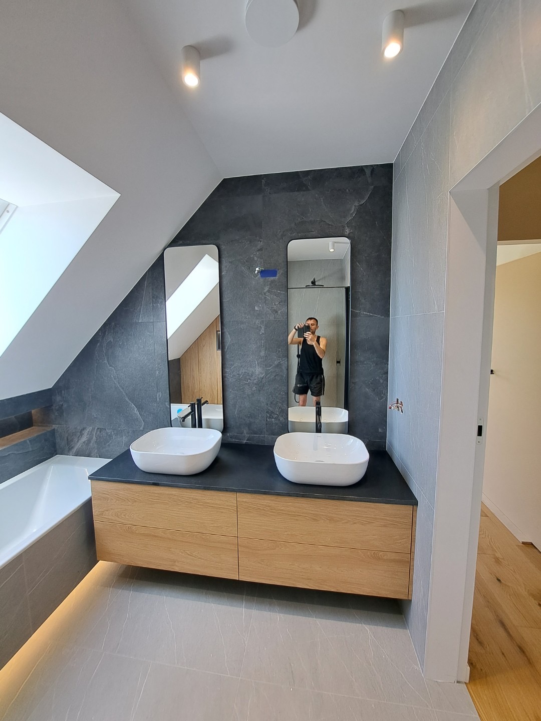 Nowoczesna łazienka na poddaszu z dwoma umywalkami nablatowymi, drewnianą szafką, szarą glazurą imitującą kamień i podświetleniem LED pod wanną. W lustrze odbija się osoba robiąca zdjęcie.