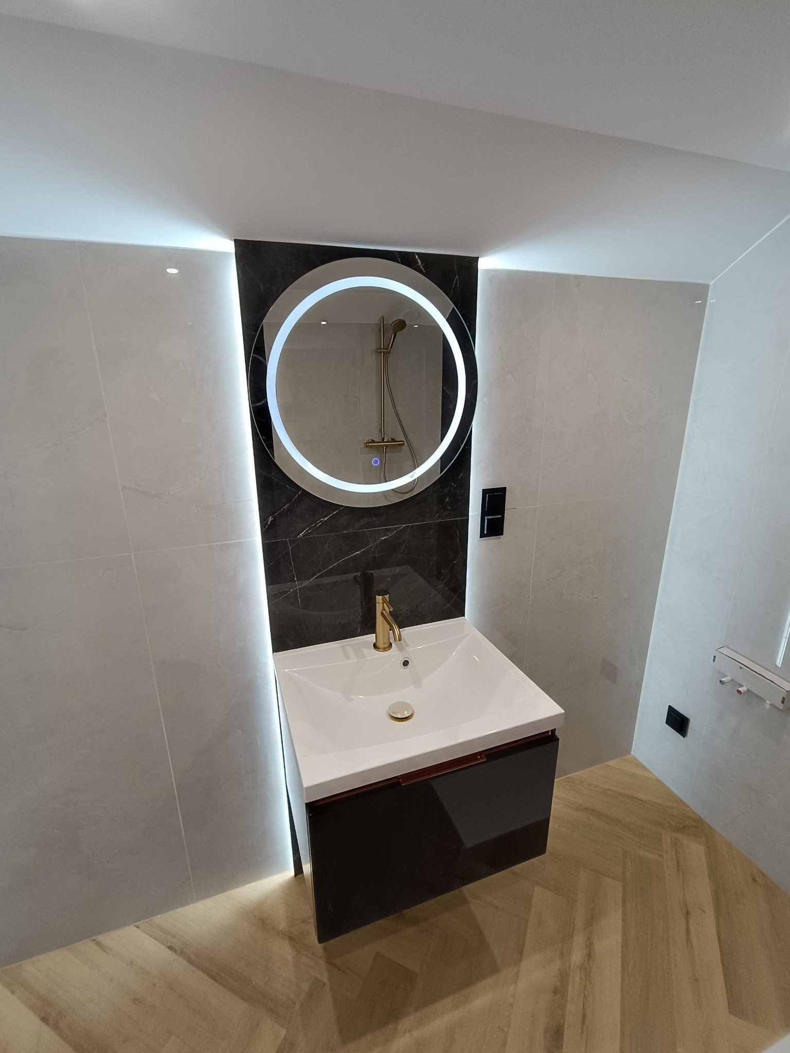 Nowoczesna łazienka z okrągłym lustrem LED, białą umywalką na szafce i złotą armaturą. Ściany wyłożone jasnymi płytkami, podłoga w jodełkę. Minimalistyczny design.