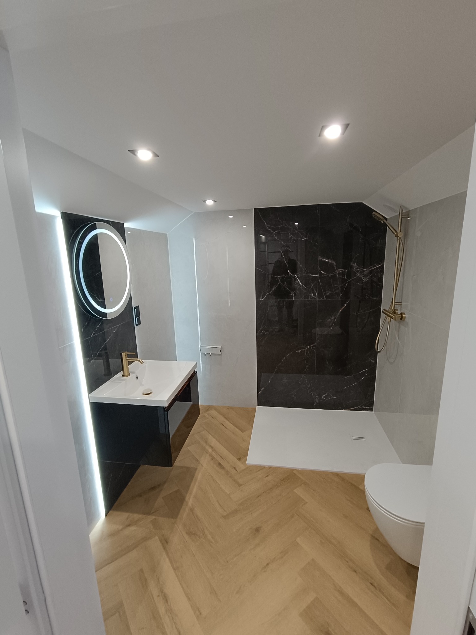 Nowoczesna łazienka z czarnymi płytkami imitującymi marmur, drewnianą podłogą w jodełkę i okrągłym lustrem z podświetleniem LED. Złota armatura dodaje elegancji.