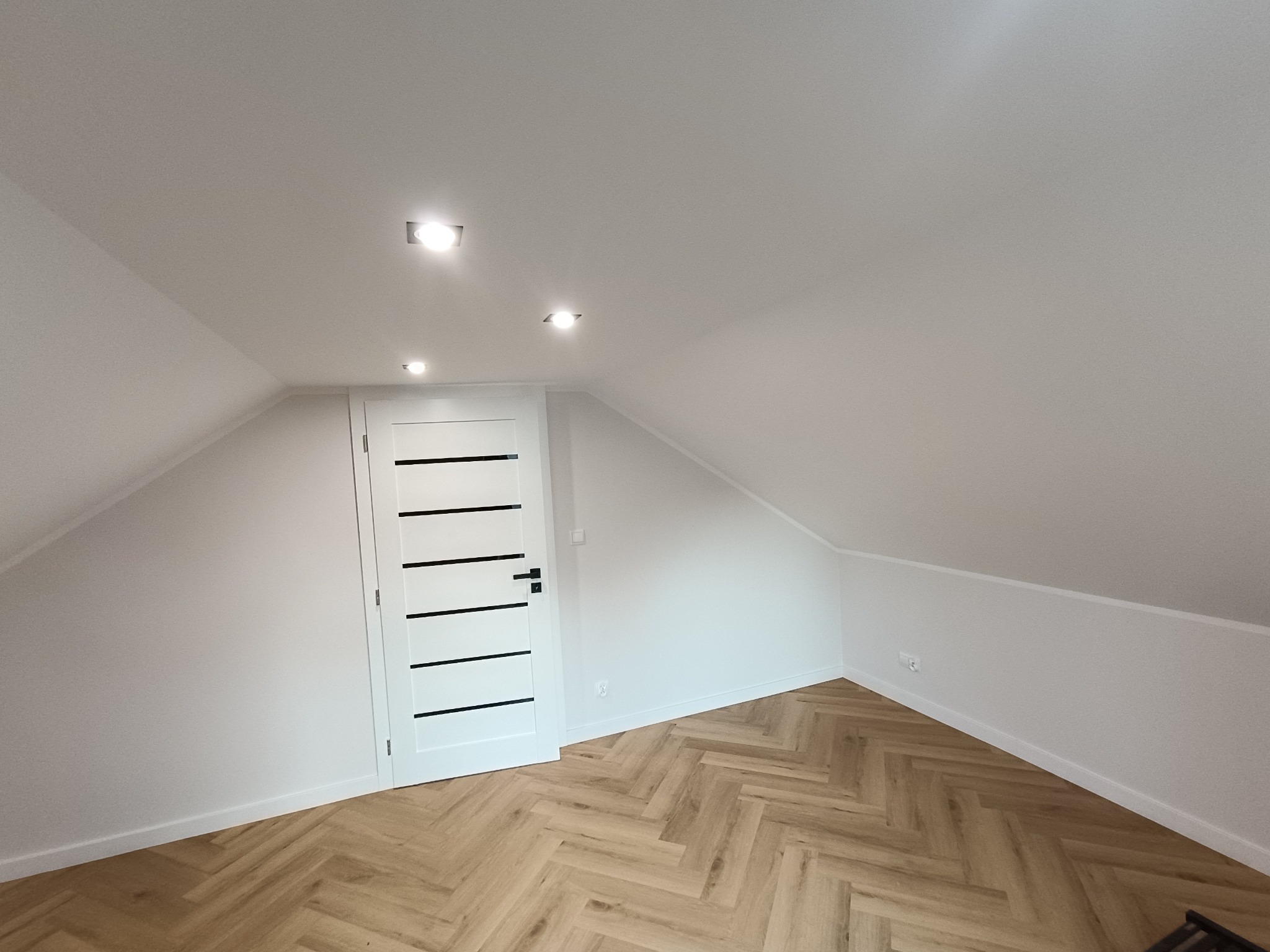 Jasne poddasze po remoncie: białe ściany, parkiet w jodełkę, nowoczesne drzwi z czarnymi akcentami i punktowe oświetlenie sufitowe. Minimalistyczny design.