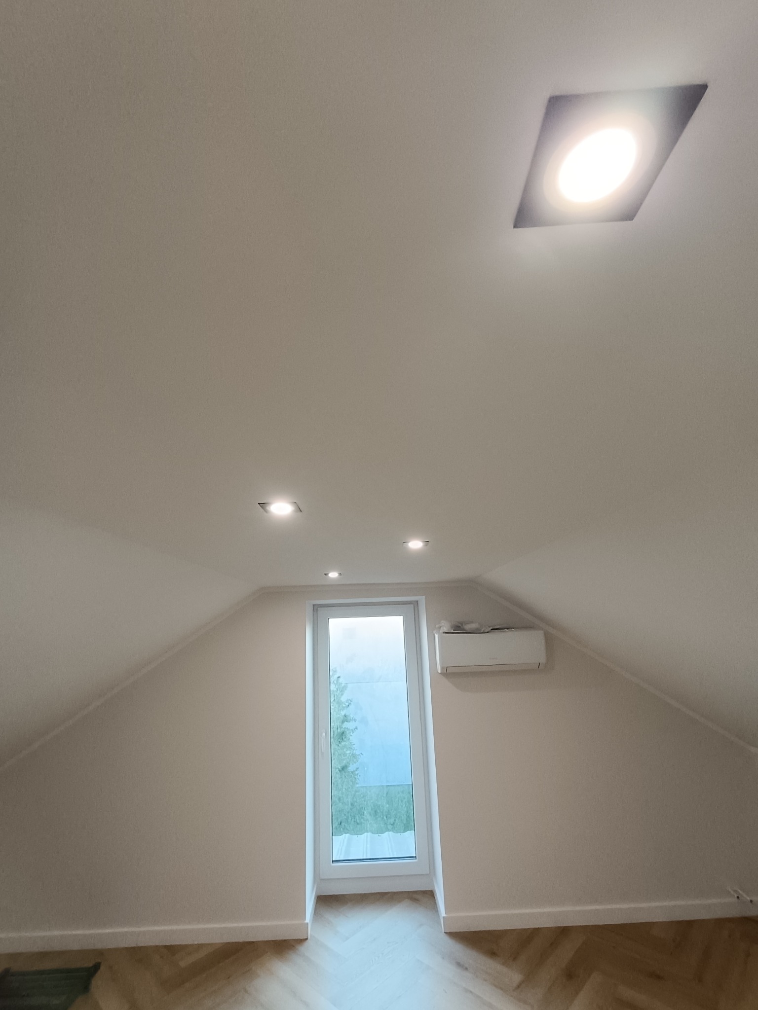 Poddasze po remoncie: skośne ściany w jasnym odcieniu, parkiet w jodełkę, okno z widokiem na zieleń, klimatyzator i nowoczesne oświetlenie sufitowe.