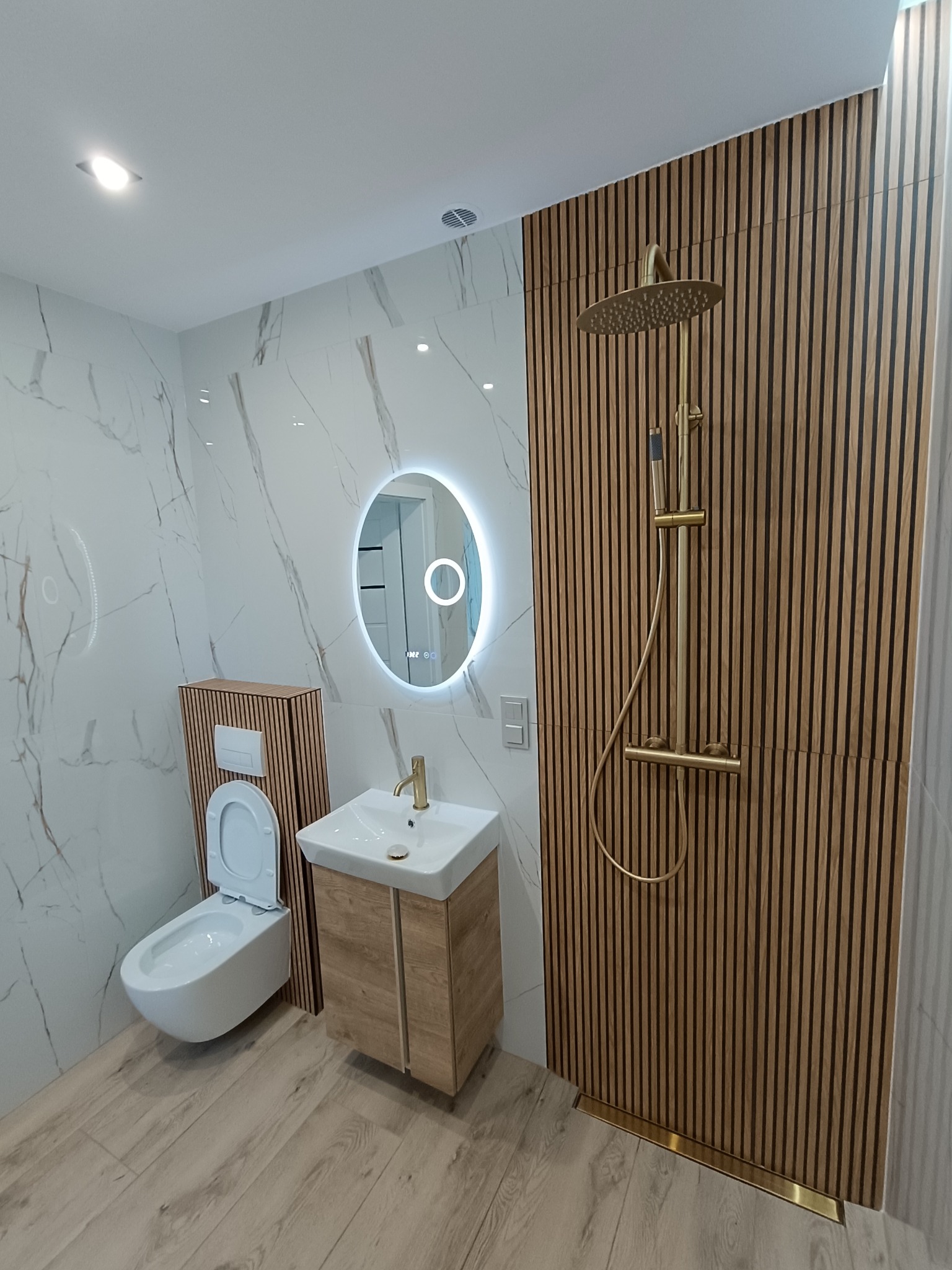 Nowoczesna łazienka z marmurowymi płytkami, drewnianymi lamelami, okrągłym lustrem LED i złotą armaturą. Uwagę przykuwa minimalistyczny design i dbałość o detale.