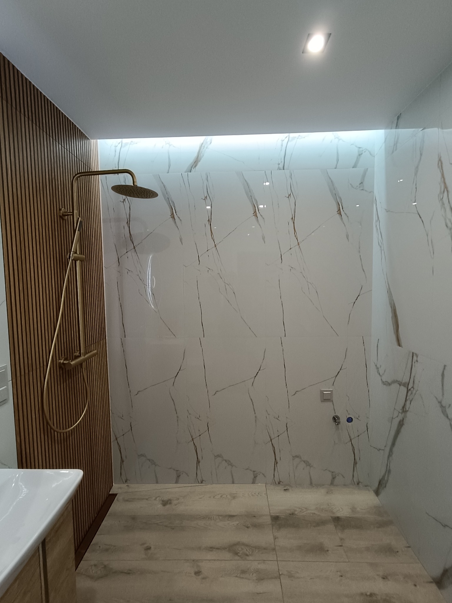 Nowoczesna kabina prysznicowa z panelami drewnianymi i marmurowymi płytkami. Złota armatura, subtelne oświetlenie LED. Minimalistyczny design, jasne kolory.