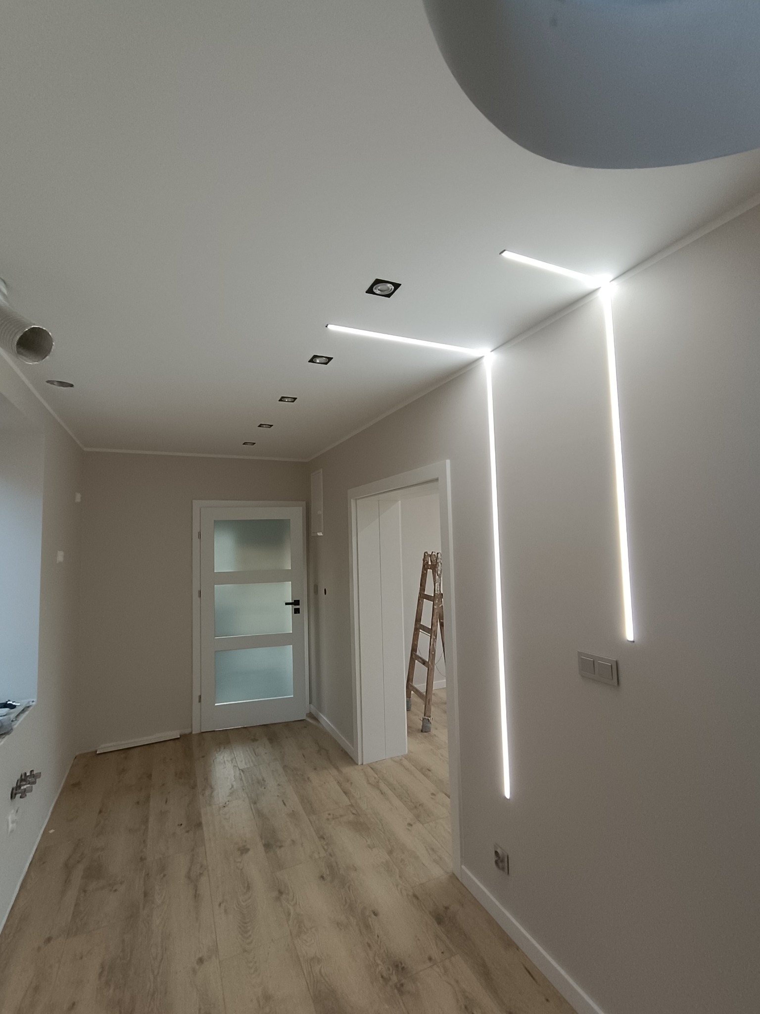 Nowoczesne oświetlenie LED w wykończonym wnętrzu: pionowe linie światła na ścianie, drewnopodobna podłoga, białe drzwi i sufit z elementami dekoracyjnymi.