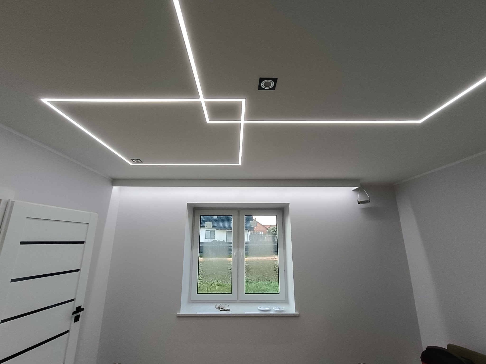 Nowoczesne oświetlenie sufitowe LED w kształcie geometrycznym, z widocznym oknem i drzwiami w minimalistycznym wnętrzu. Białe ściany i sufit.