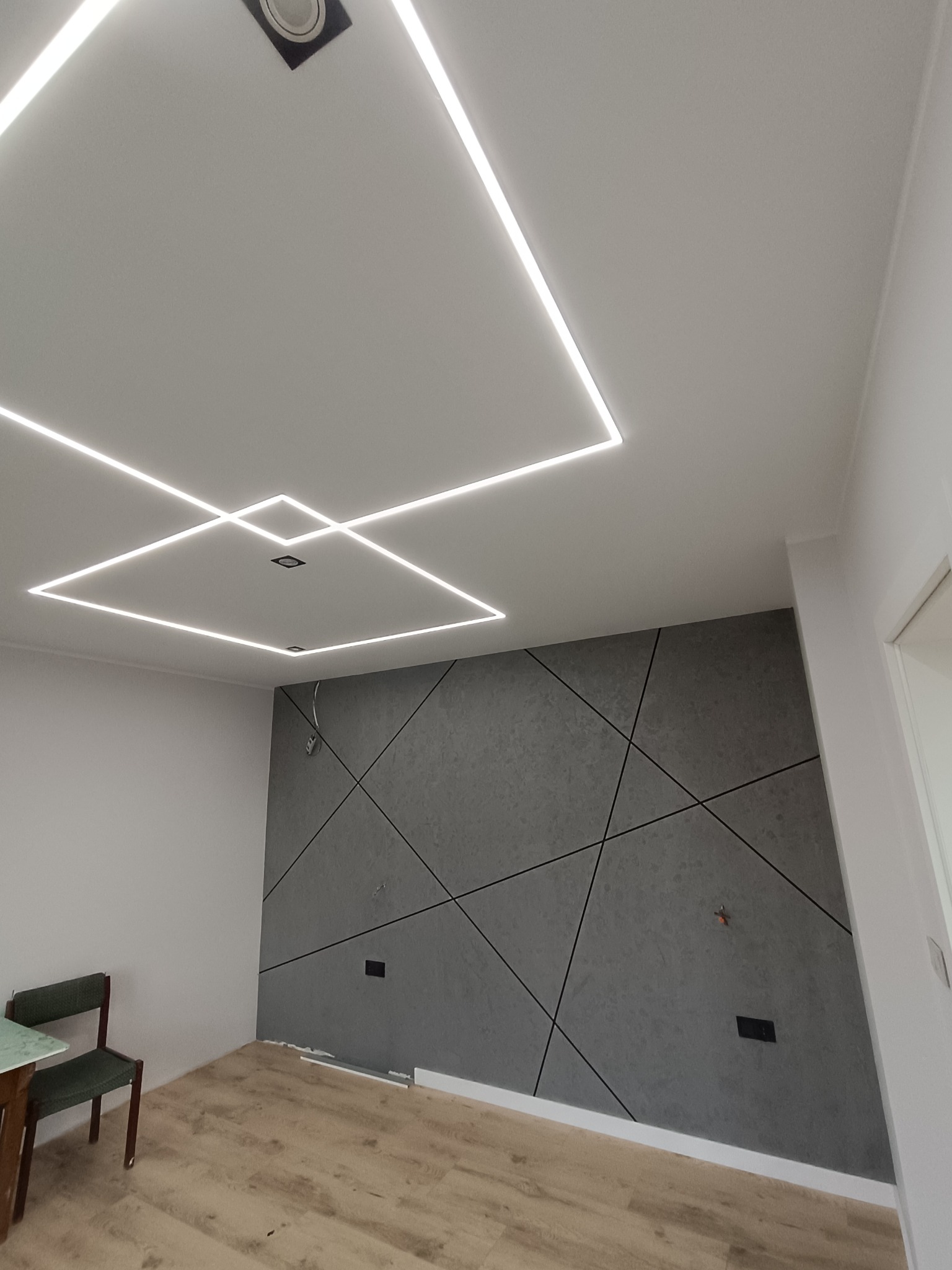 Nowoczesne wnętrze: szara ściana z geometrycznym wzorem, podświetlany sufit z kwadratowymi lampami LED, drewniana podłoga i krzesło w rogu pokoju.