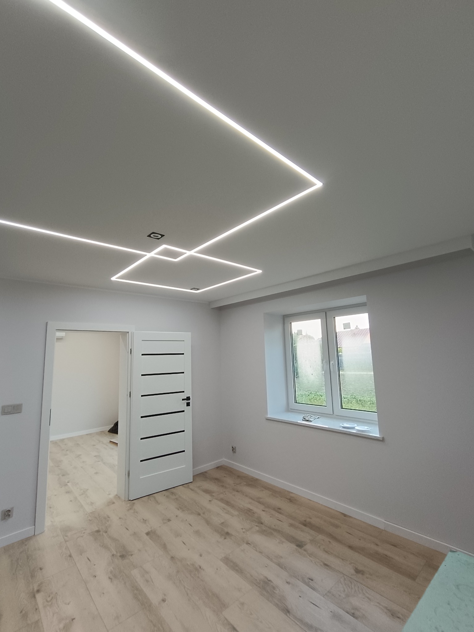 Jasne wnętrze pokoju po remoncie: drewniana podłoga, białe ściany, okno i drzwi z czarnymi akcentami. Na suficie geometryczne oświetlenie LED.