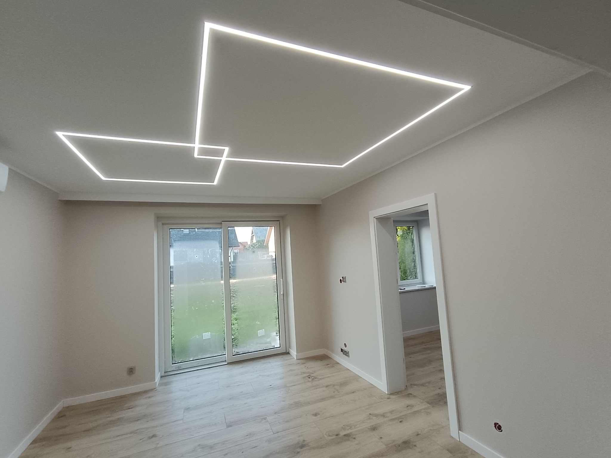 Nowoczesne oświetlenie sufitowe LED w kształcie geometrycznym w jasnym, odnowionym wnętrzu z drewnianą podłogą i dużym oknem balkonowym.