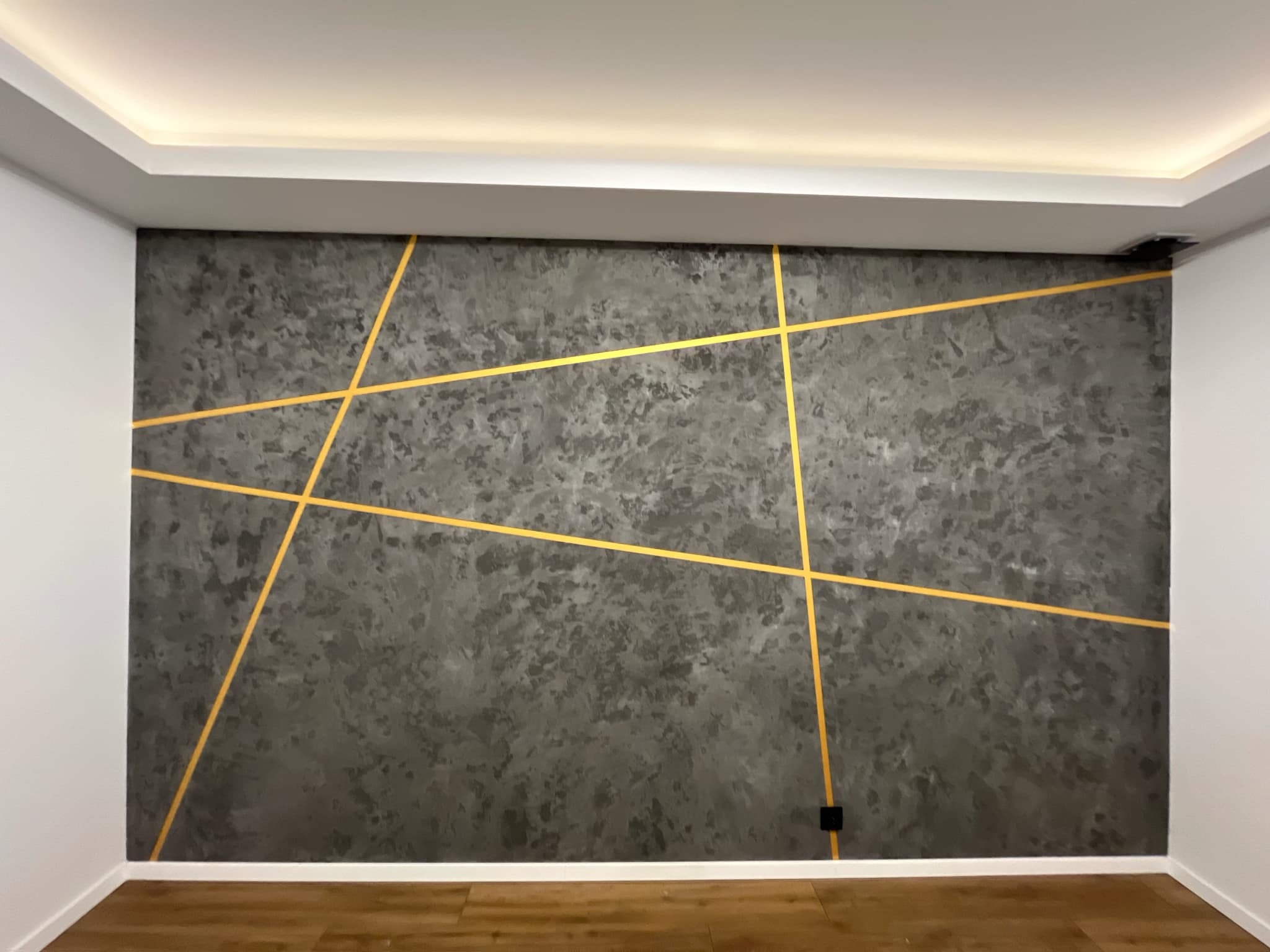 Ściana z efektem dekoracyjnym w odcieniach szarości, przecięta złotymi liniami. Widoczne drewniane panele podłogowe i białe ściany boczne. Nowoczesny design wnętrza.