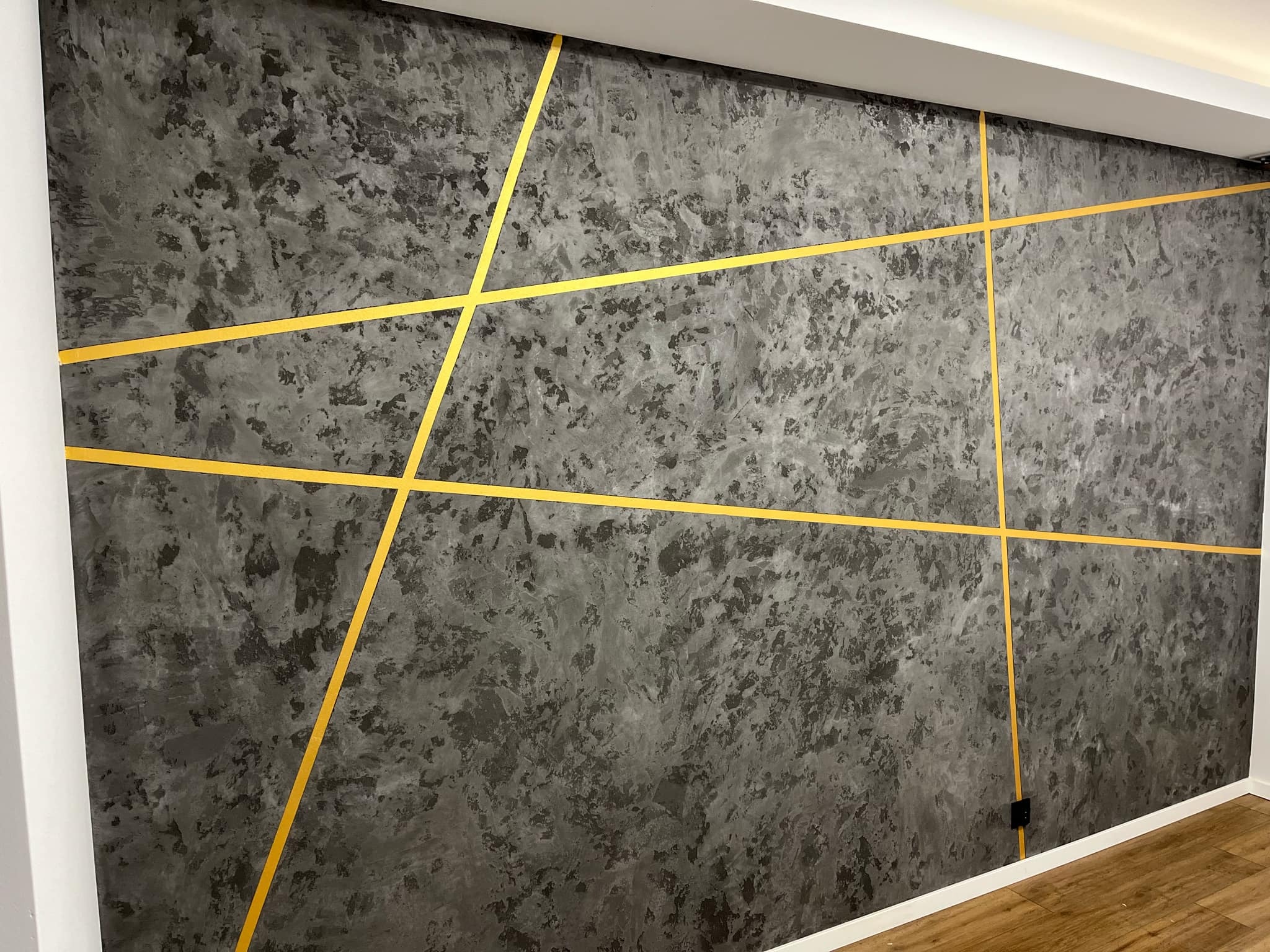 Ściana z efektem dekoracyjnym w odcieniach szarości, przecięta złotymi, geometrycznymi liniami. Nowoczesny design wnętrza z widocznym gniazdkiem elektrycznym.