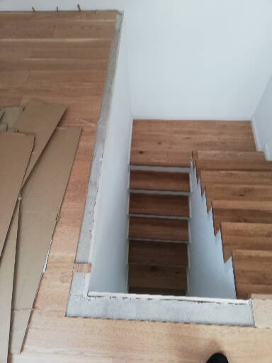 Montaż drewnianych schodów: widok z góry na schody w trakcie układania, widoczne kartony zabezpieczające podłogę i fragment buta w rogu kadru.