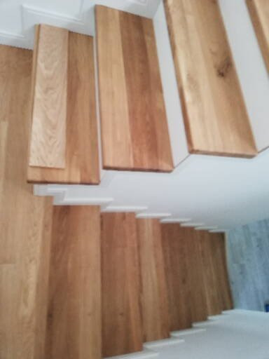 Widok z góry na jasne, drewniane schody z białymi podstopnicami, oświetlone naturalnym światłem, z delikatnym wzorem drewna widocznym na stopniach.