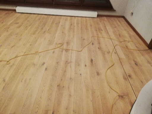 Jasna, świeżo położona podłoga z paneli z widocznym kablem elektrycznym na powierzchni, częściowo widoczny element maszyny szlifującej w prawym dolnym rogu.