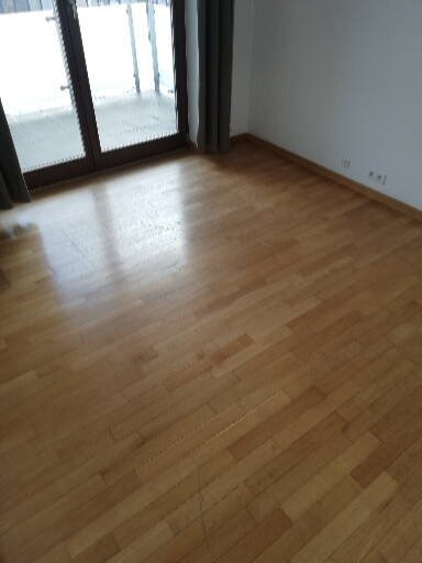 Wycyklinowana i polakierowana drewniana podłoga w pustym pokoju z oknem balkonowym i zasłonami.