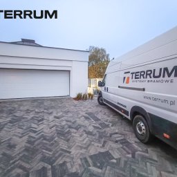 TERRUM Sp. z o.o. - Biała segmentowa brama garażowa Hormann w nowoczesnym garażu, obok zaparkowany biały samochód dostawczy z logo firmy Terrum Systemy Bramowe na tle kostki brukowej w jodełkę.