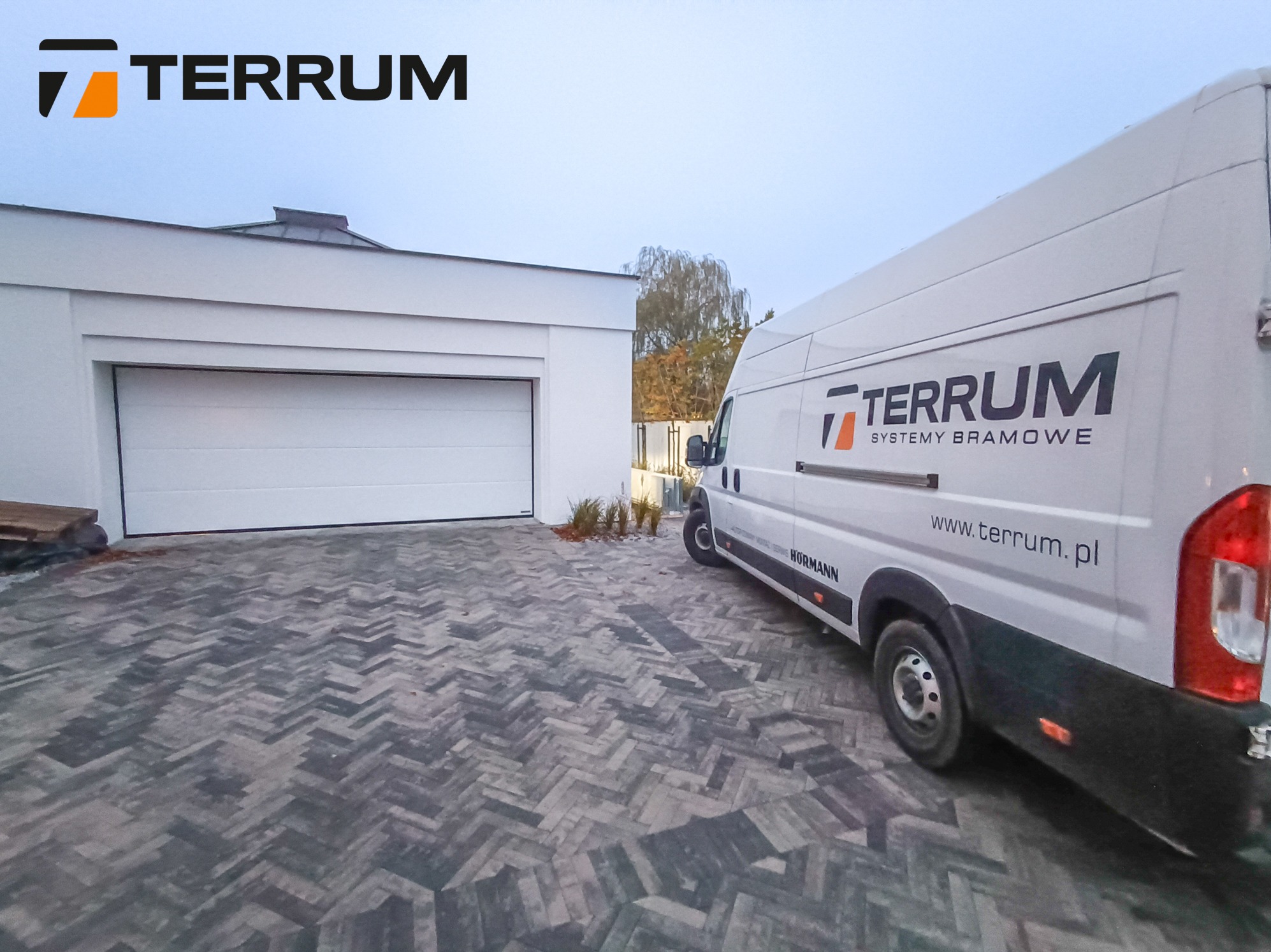 Biała segmentowa brama garażowa Hormann w nowoczesnym garażu, obok zaparkowany biały samochód dostawczy z logo firmy Terrum Systemy Bramowe na tle kostki brukowej w jodełkę.