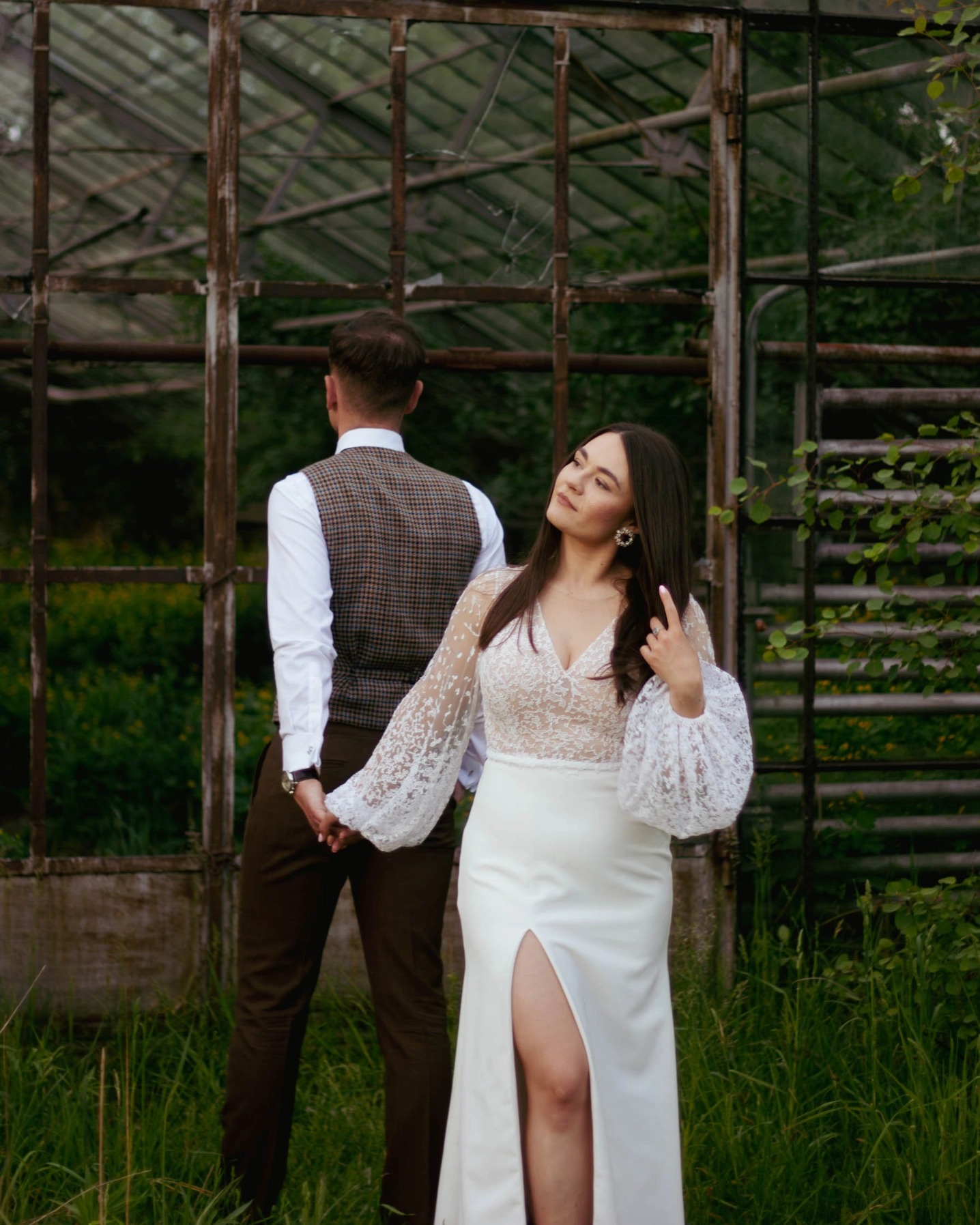 Para trzymająca się za ręce przed szklarnią: kobieta w białej sukni z koronkową górą i rozcięciem, mężczyzna w brązowej kamizelce w kratę i ciemnych spodniach, oboje patrzą w tym samym kierunku.