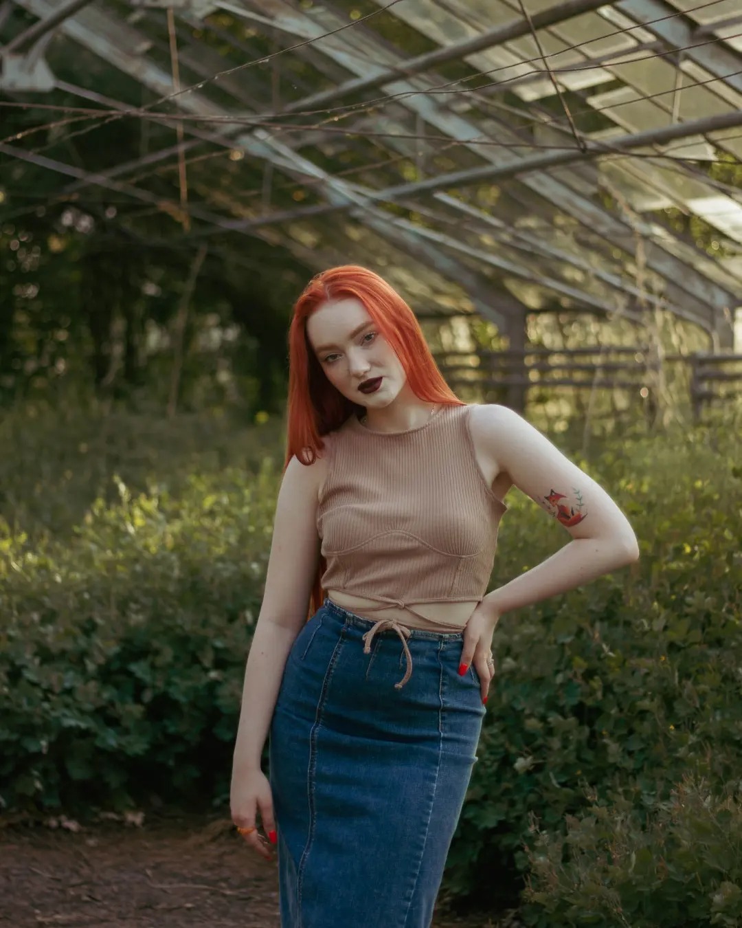 Portret kobiety z rudymi włosami w dżinsowej spódnicy i topie na tle szklarni, z tatuażem lisa na ramieniu.