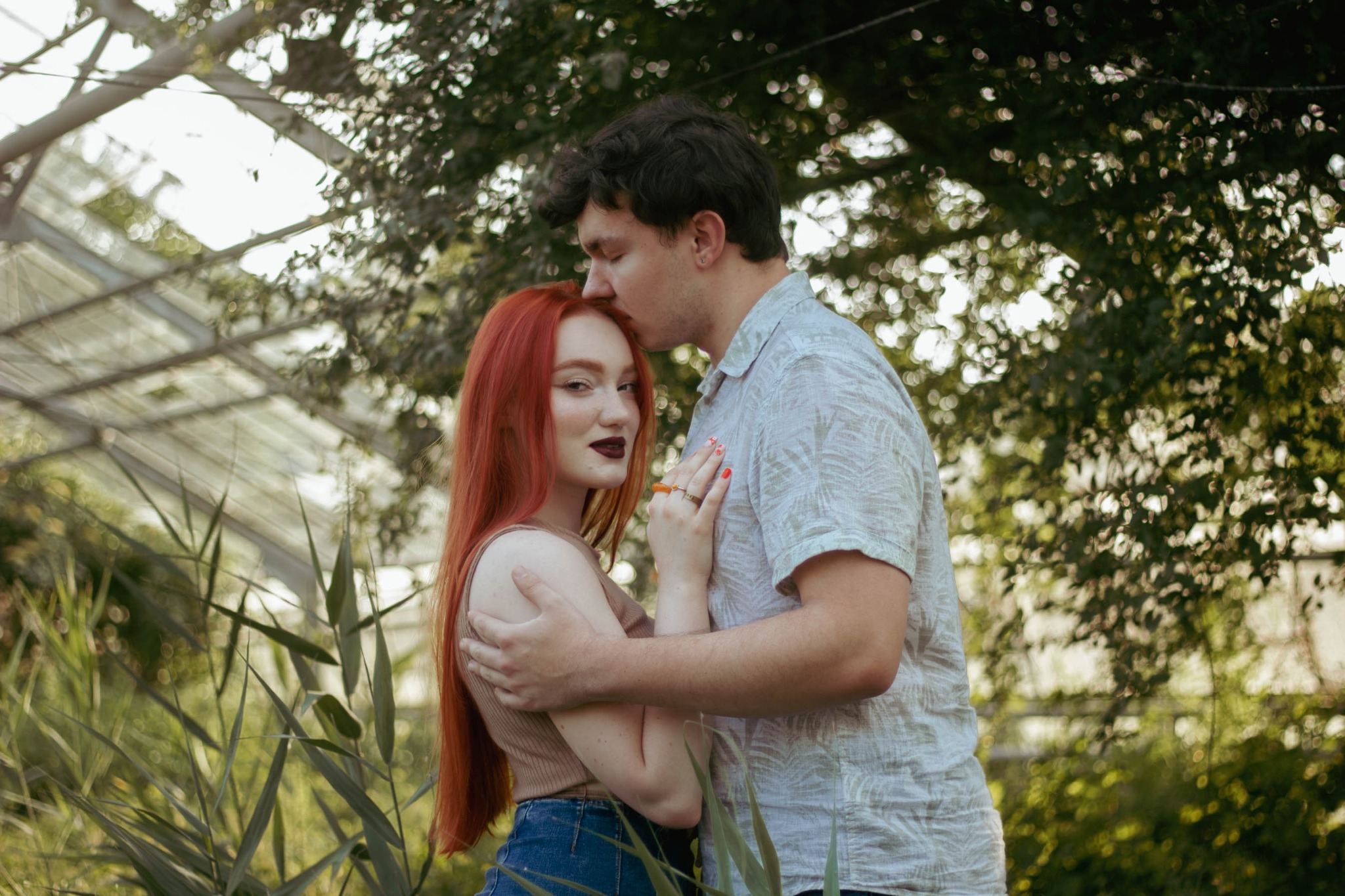 Zakochana para w szklarni: mężczyzna całuje kobietę w czoło, ona patrzy w obiektyw z delikatnym uśmiechem, w tle bujna roślinność i konstrukcja szklarni.