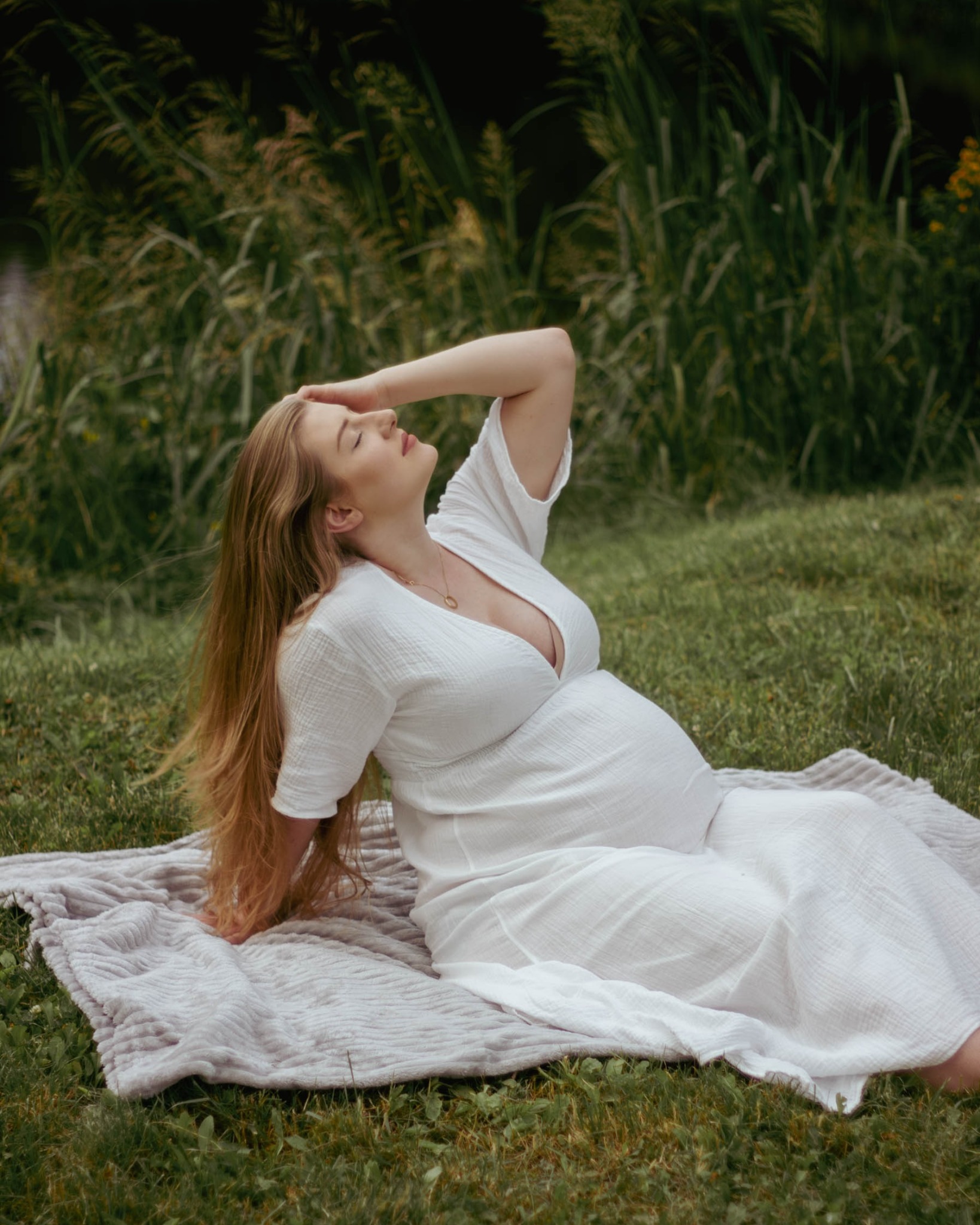 Kobieta w białej sukience, leżąca na kocu na trawie, z ręką uniesioną do czoła, w tle zieleń i woda.