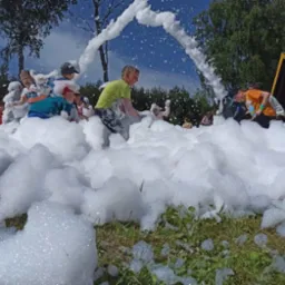 Letnia zabawa w pianie dla dzieci na świeżym powietrzu. Grupa dzieci brodzi i bawi się w dużej ilości białej piany, z drzewami i fragmentem budynku w tle.