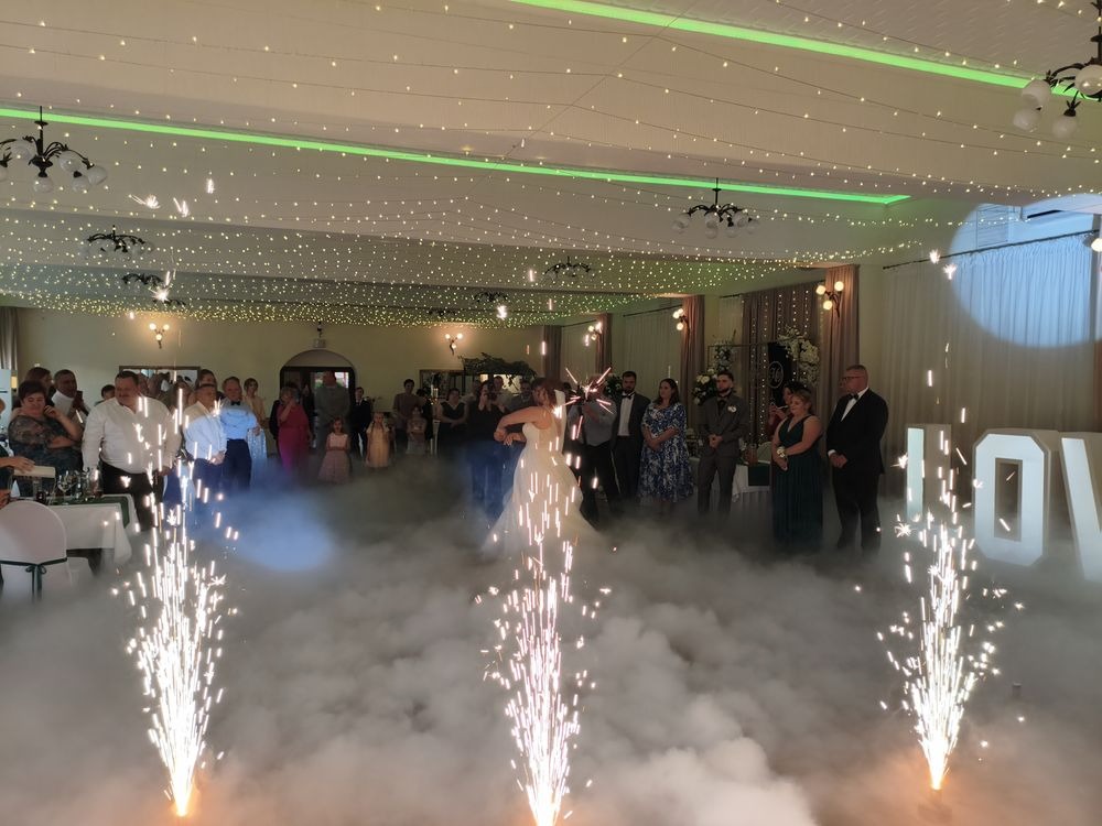 Para młoda tańczy pierwszy taniec w chmurze dymu i blasku fontann iskier na tle gości i dekoracji sali weselnej. Napis LOVE w tle.