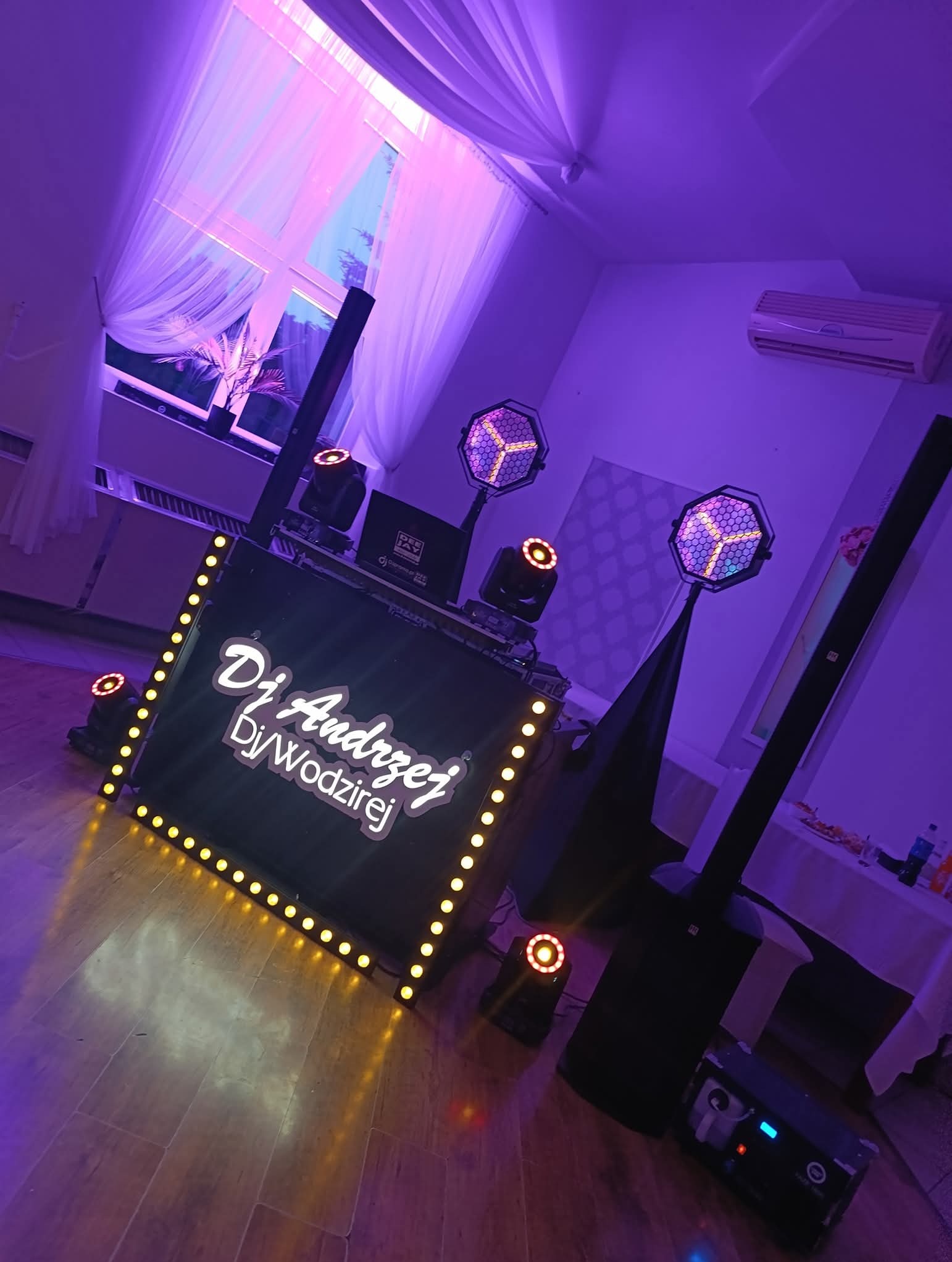Oświetlona konsola DJ'a 'DJ Andrzej DJ Wodzirej' z nagłośnieniem i efektami świetlnymi w fioletowym oświetleniu sali. Widoczne okno z firanami.