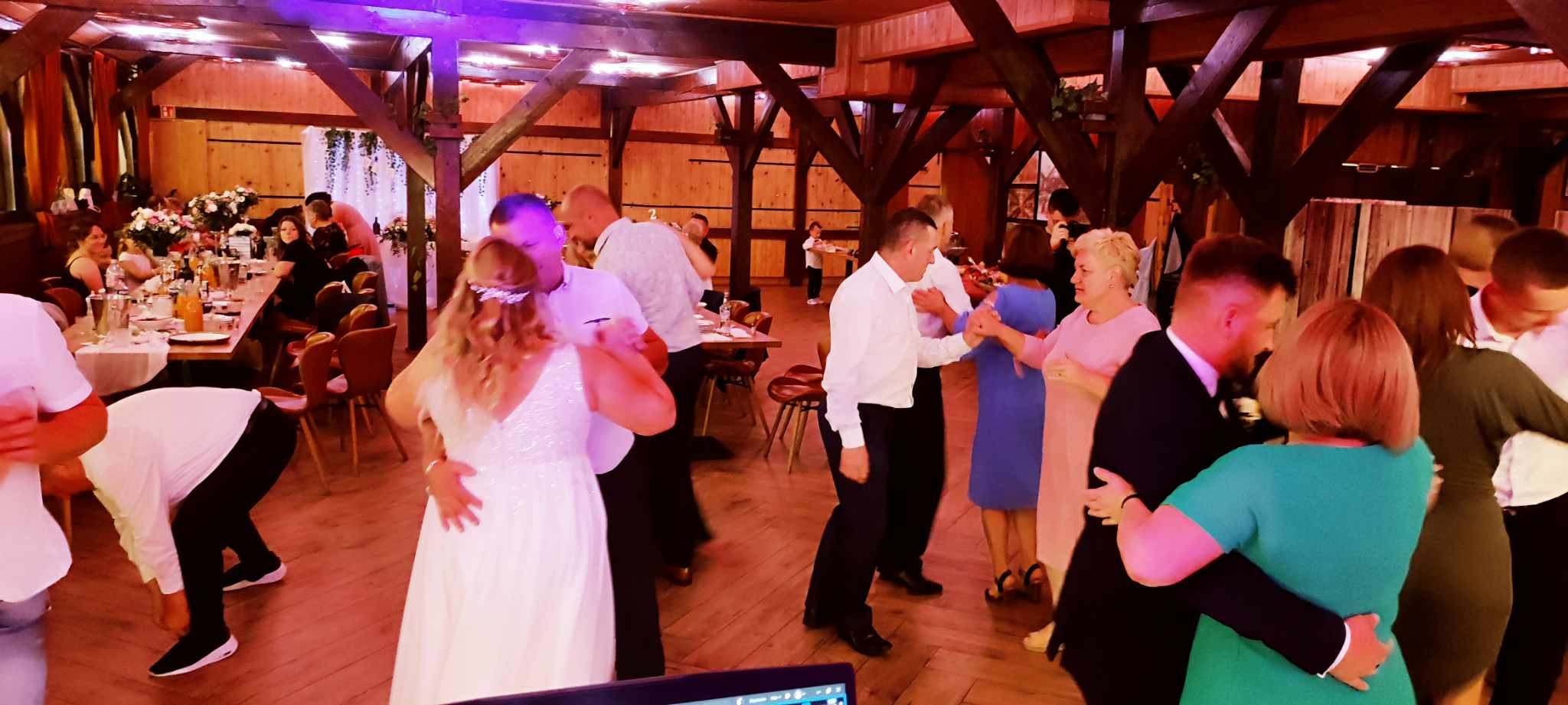 Goście weselni tańczą na parkiecie w drewnianej sali balowej, widoczne nakryte stoły i dekoracje kwiatowe.