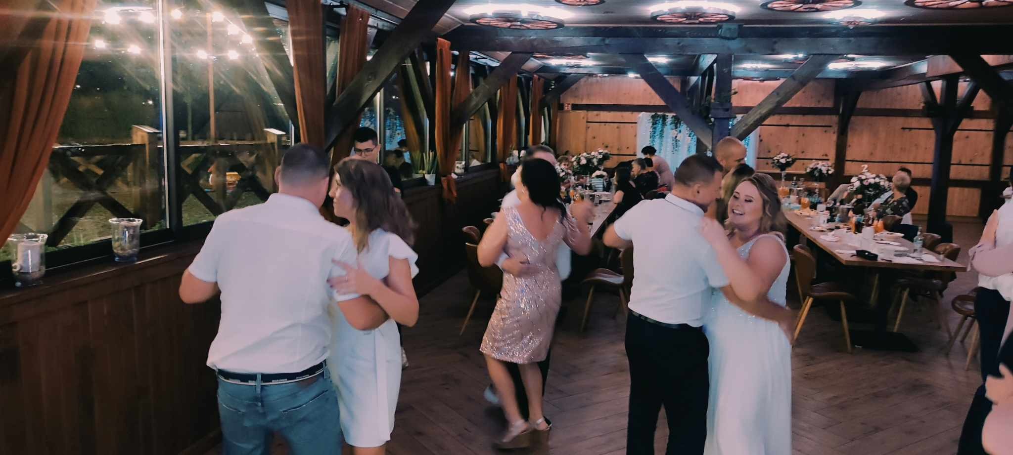 Goście weselni tańczą w rustykalnej sali z drewnianą konstrukcją dachu, dekoracjami kwiatowymi na stołach i oświetleniem w tle.