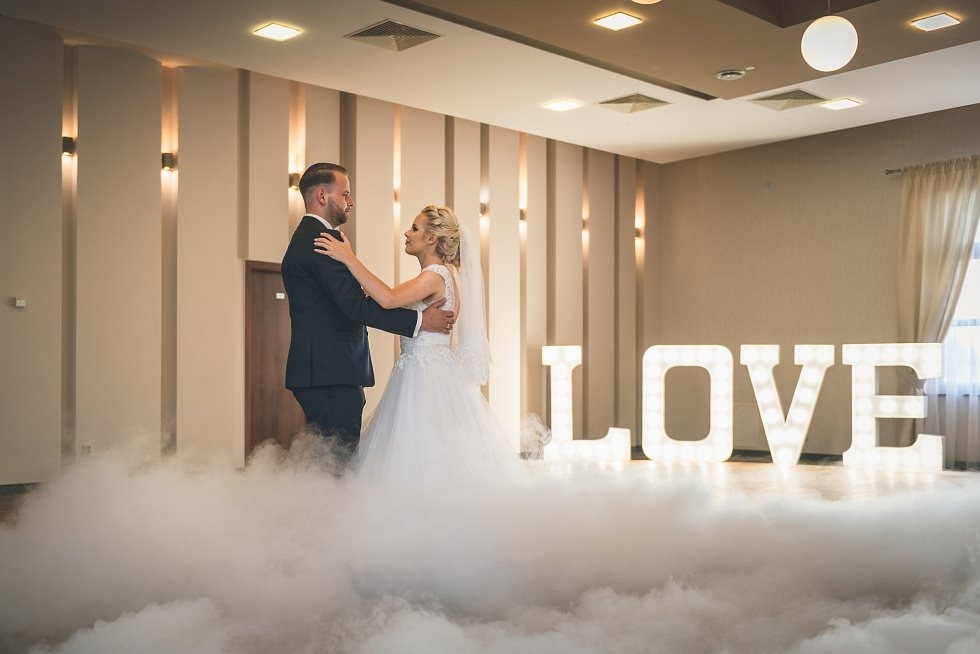 Para młoda tańczy w chmurze dymu na tle podświetlonego napisu LOVE w sali weselnej.