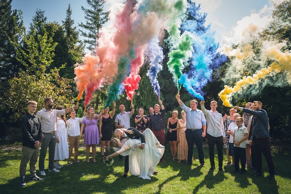 Para młoda w objęciach na tle grupy osób z kolorowymi racami dymnymi na zielonej trawie, drzewa w tle.