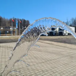 Strumień wody z niebieskiej rury na tle brukowanej powierzchni, z widocznymi budynkami, drzewami i flagami w oddali.