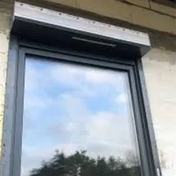 Zamknięta, szara osłona okienna z widocznym mechanizmem zwijania, zamontowana na oknie z refleksami nieba i drzew w szybie, na tle surowej ściany budynku.