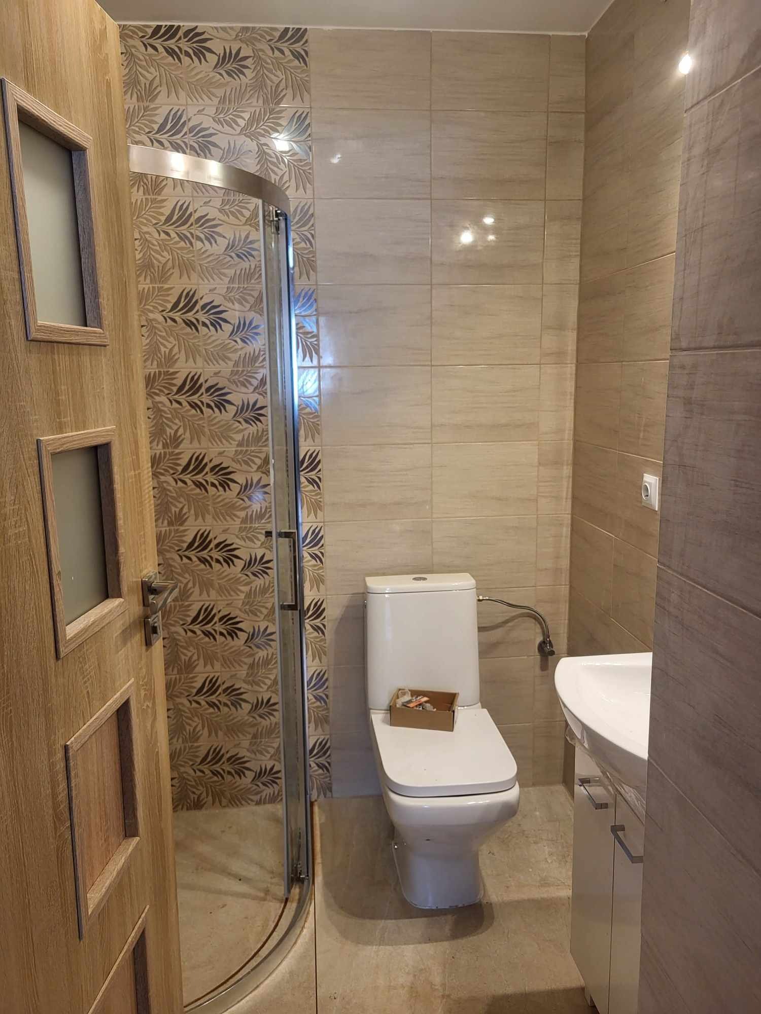 Ukończona łazienka w beżowych odcieniach z kabiną prysznicową ze wzorem liściastym na ścianie, toaleta z pudełkiem na desce i umywalka z szafką, widziana z perspektywy lekko otwartych, drewnianych...