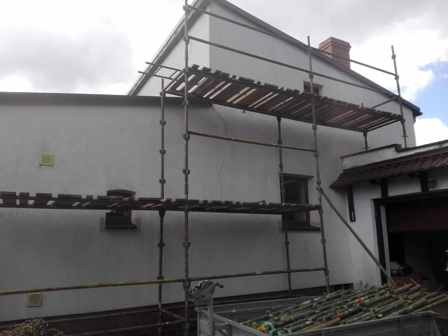 Dom jednorodzinny z białymi ścianami, częściowo zakryty rusztowaniem podczas prac remontowych; widoczne okna i komin z czerwonej cegły.