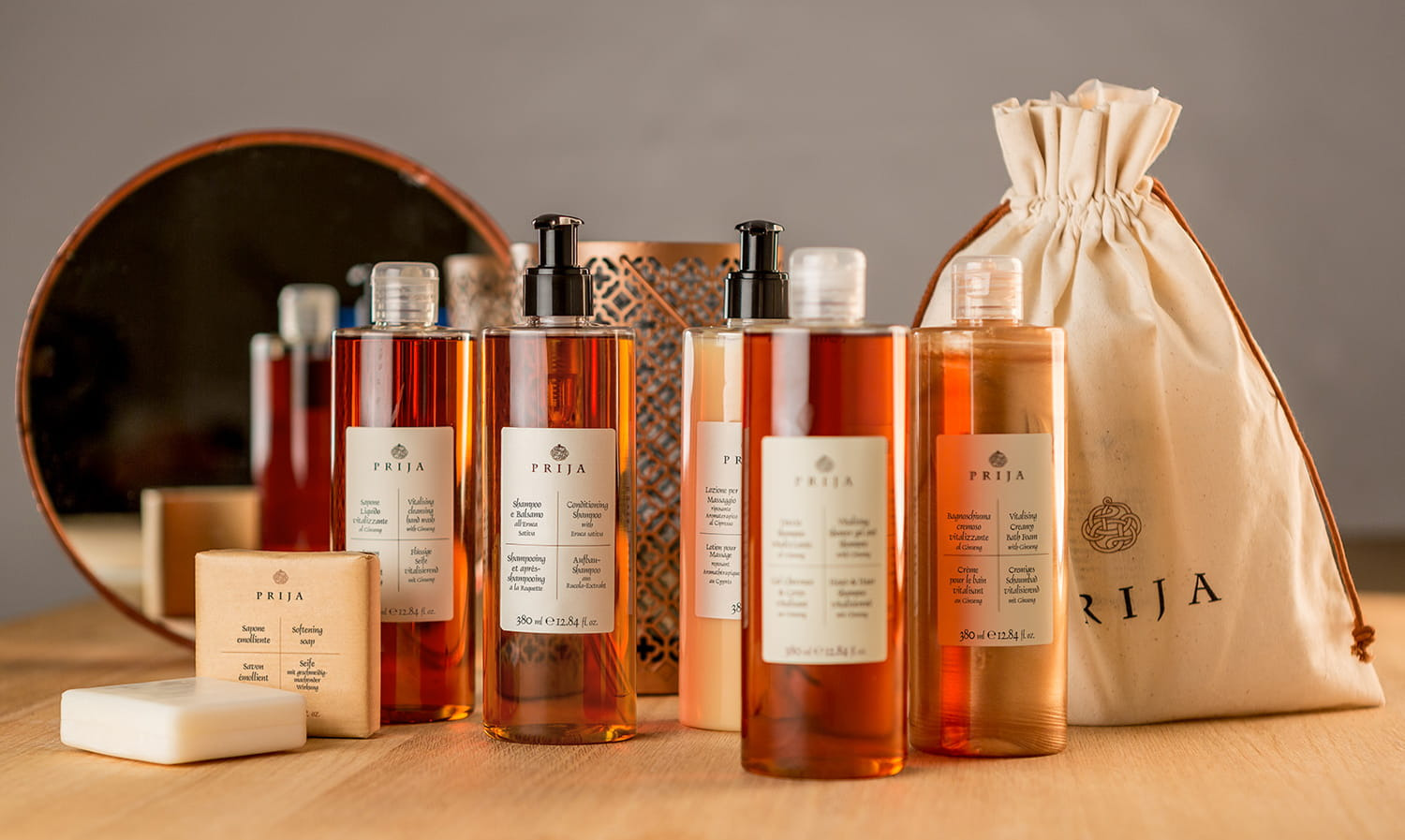 Zestaw kosmetyków Prija na drewnianym blacie: mydło w kostce, butelki z szamponem i żelem pod prysznic o bursztynowej barwie, woreczek z logo, odbicie w okrągłym lustrze w tle.
