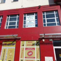 Fasada budynku z czerwonymi panelami, na której widnieje szyld 'Biuro Rachunkowe DwaPlusCztery' oraz numer telefonu, poniżej sklep z odzieżą dziecięcą 'U Sowy'.