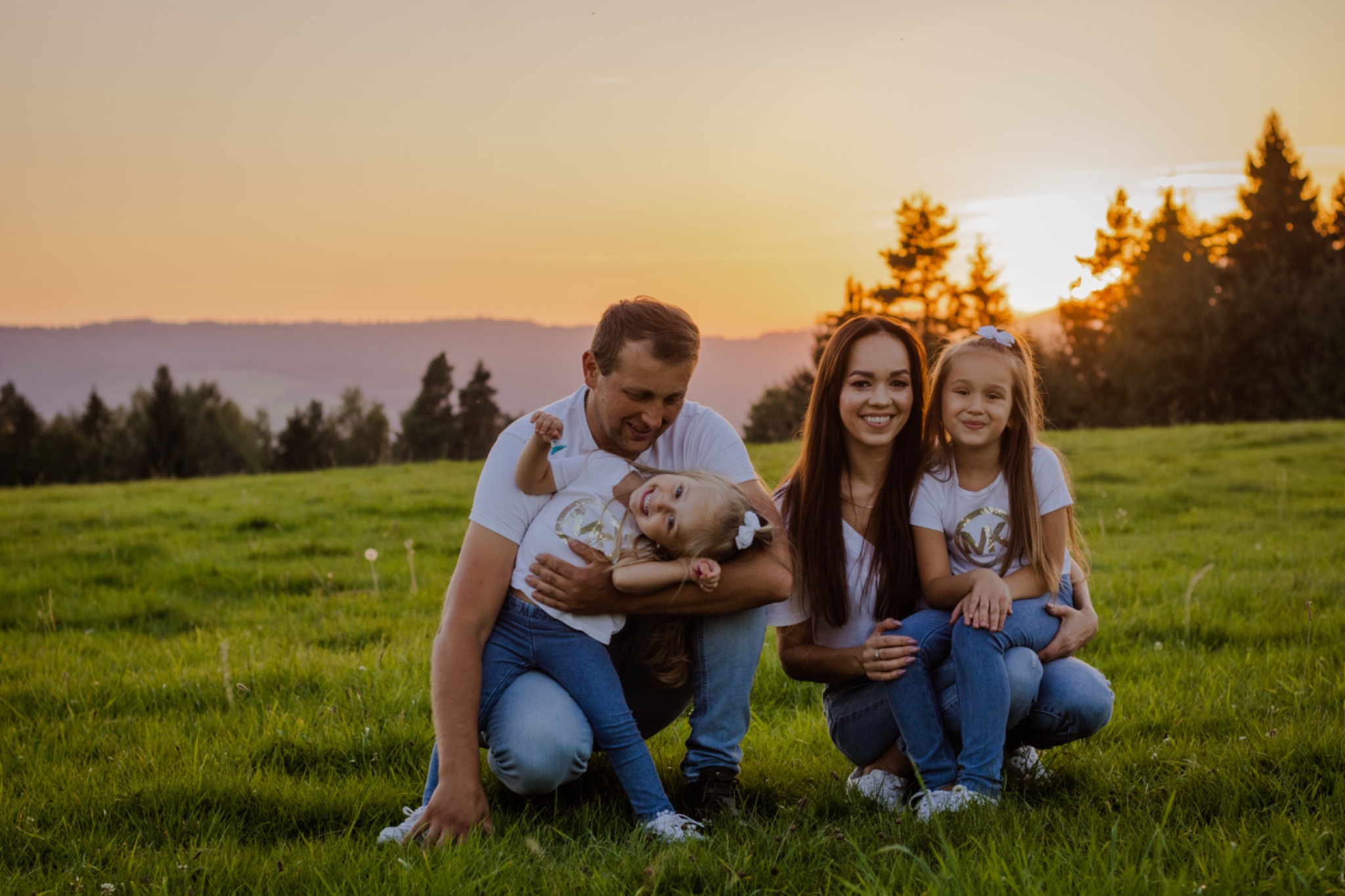 Rodzinne zdjęcie w plenerze: rodzice z dwiema córkami siedzą na trawie na tle zachodzącego słońca i lasu.