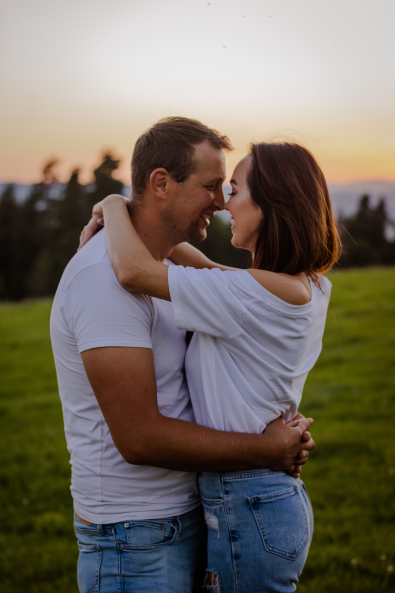 Para zakochanych w objęciach na tle zachodzącego słońca, ubrana w białe koszulki i jeansy, z uśmiechami na twarzach.