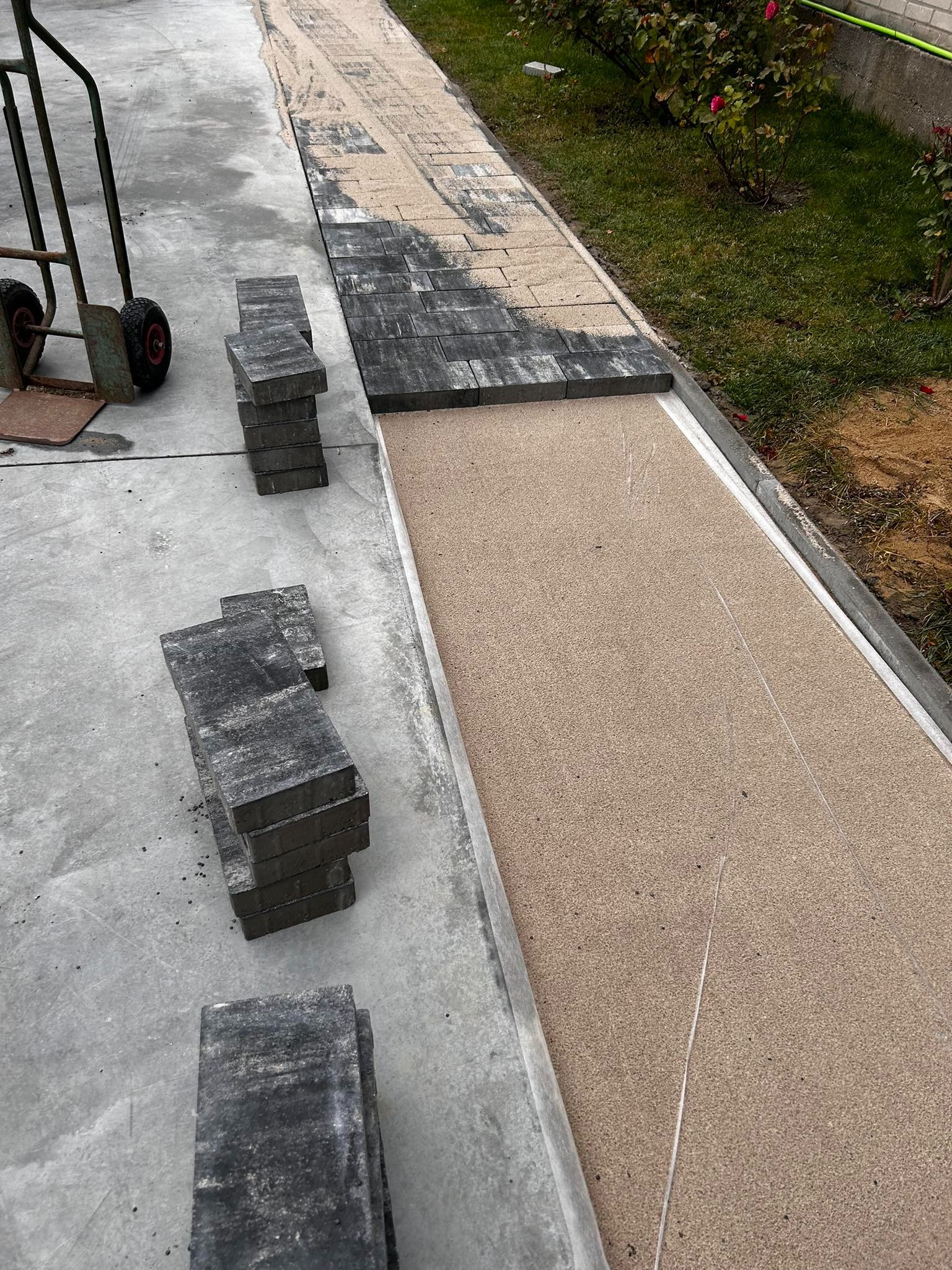 Montaż kostki brukowej na podsypce piaskowej, widoczny wózek transportowy i ułożone stosy kostek w odcieniach szarości.