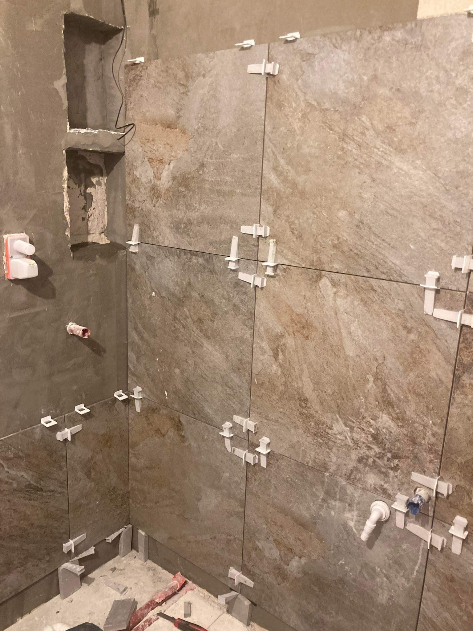 Układanie płytek imitujących kamień w rogu łazienki, widoczne białe krzyżyki dystansowe i fragment instalacji hydraulicznej.