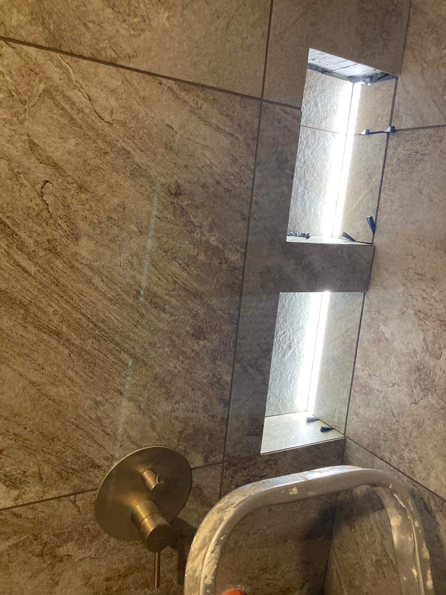 Wykończona wnęka prysznicowa z półkami wyłożona płytkami imitującymi kamień, widoczna bateria i wiadro budowlane.