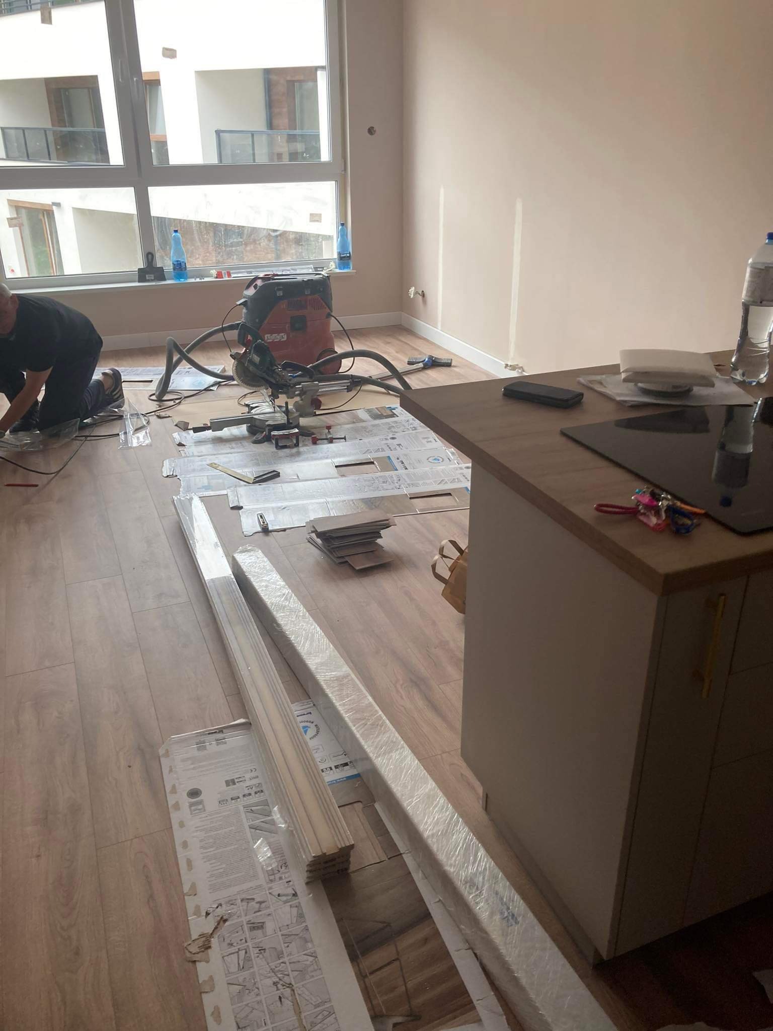 Montaż paneli podłogowych w jasnym wnętrzu, widoczny pracownik przycinający listwy przypodłogowe, odkurzacz przemysłowy i narzędzia na podłodze, zarys kuchni z płytą indukcyjną w tle.