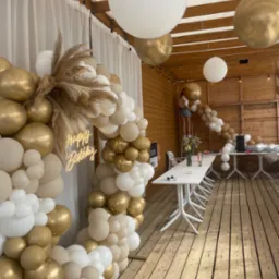 Dekoracja urodzinowa z białych, złotych i beżowych balonów, trzcin oraz neonu 'Happy Birthday' na drewnianym tarasie, osoba na drabinie mocuje girlandę balonową pod drewnianym zadaszeniem.