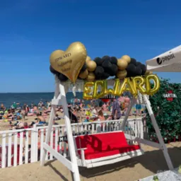 Plażowa impreza pożegnalna dla Edwarda: biała huśtawka z czerwonym siedziskiem, złote balony z napisem 'Edward Będziemy tęsknić' i 'Edward', czarno-złota girlanda balonowa, stół z napojami, tło...