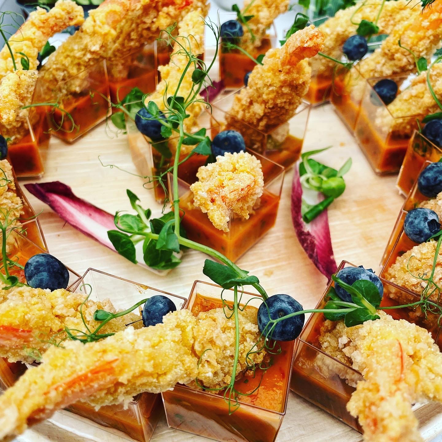 Aperitif z krewetką w tempurze, sosem pomidorowym, borówkami i mikro-ziołami, podany w małych, kwadratowych szklankach na drewnianej desce.