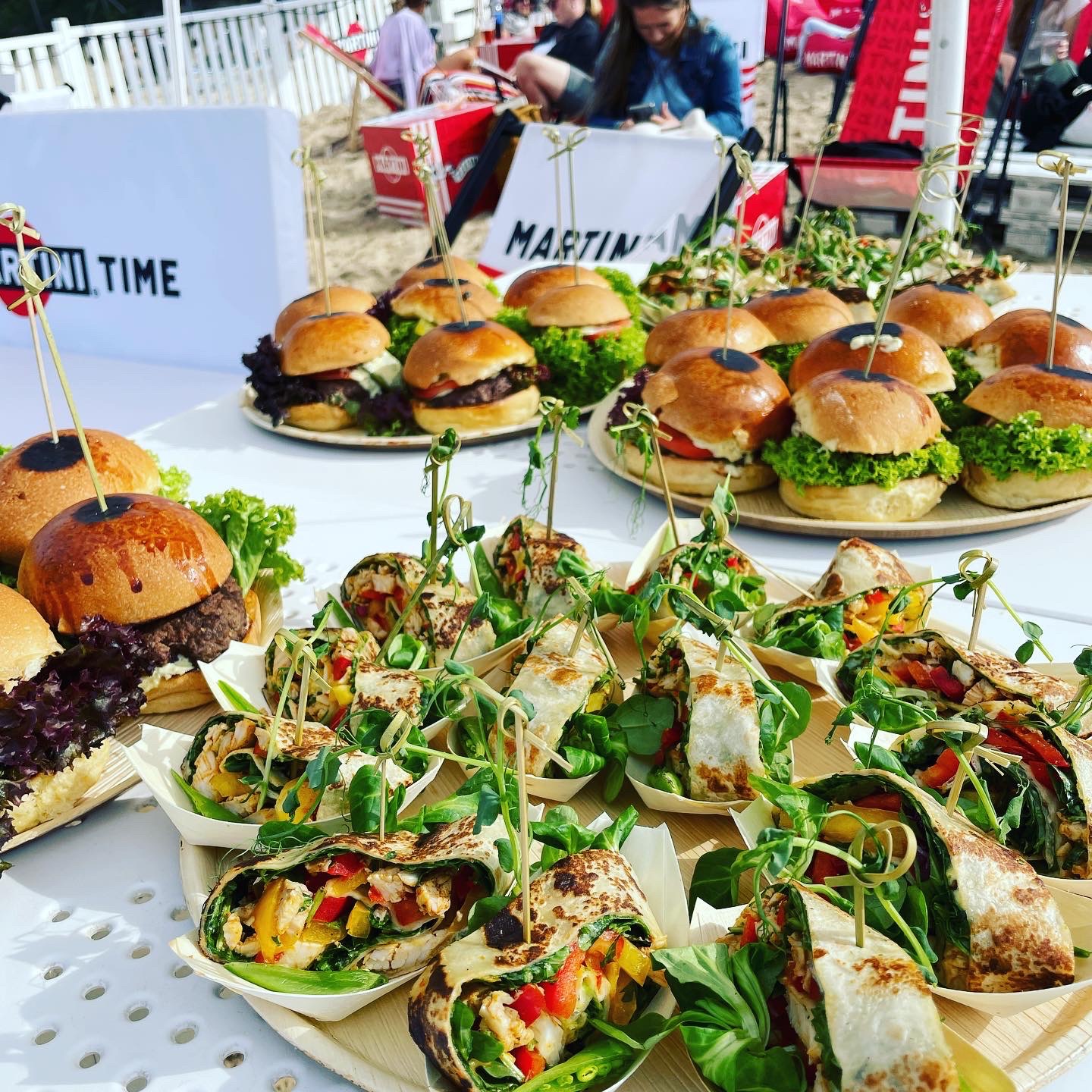 Stół z cateringiem: mini burgery i wrap-y z warzywami i kurczakiem, dekorowane wykałaczkami, z logo Martini Time w tle, na imprezie plenerowej.