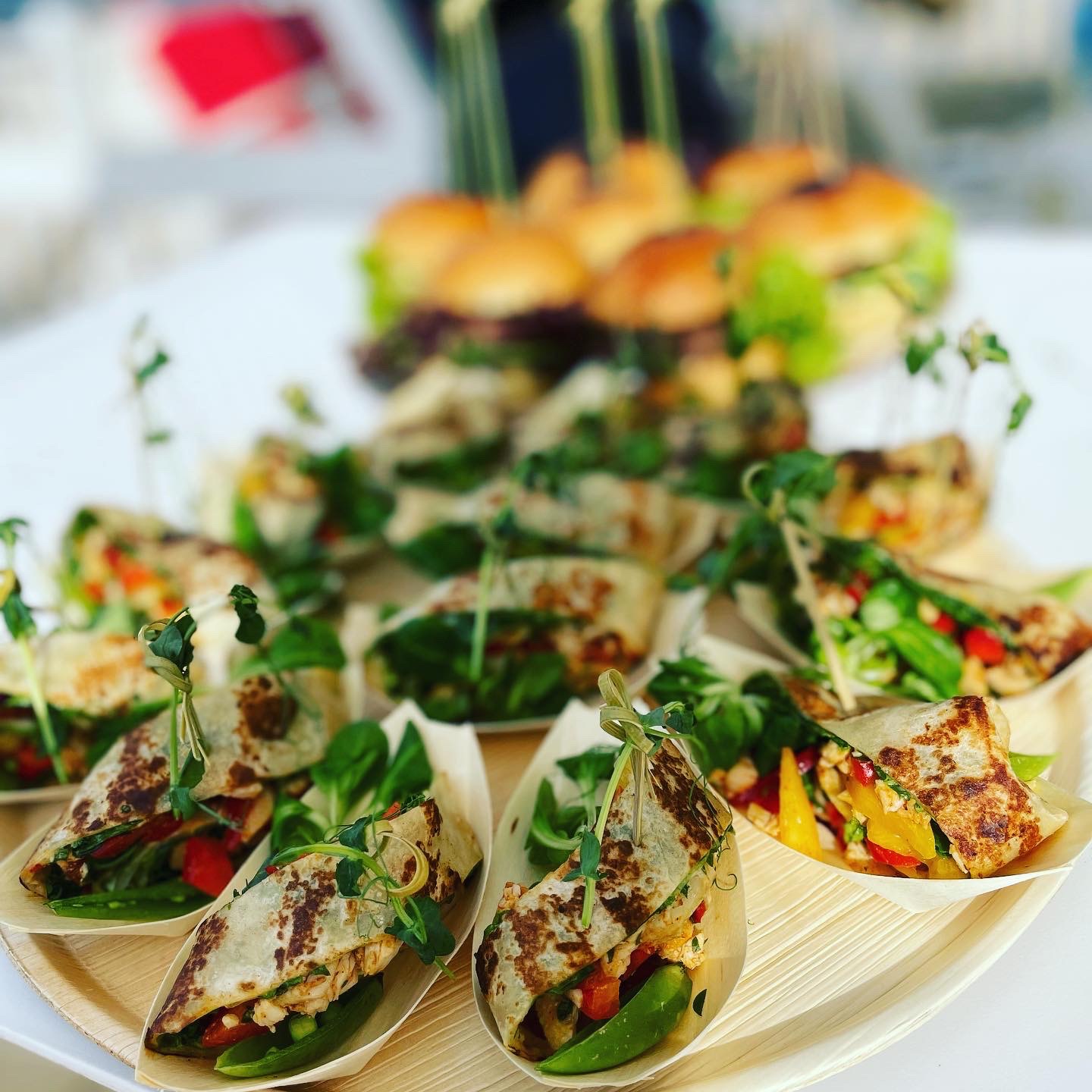 Elegancko podane przekąski: mini tortille z warzywami i mikro-ziołami w drewnianych łódeczkach oraz mini burgery w tle, catering na imprezę.
