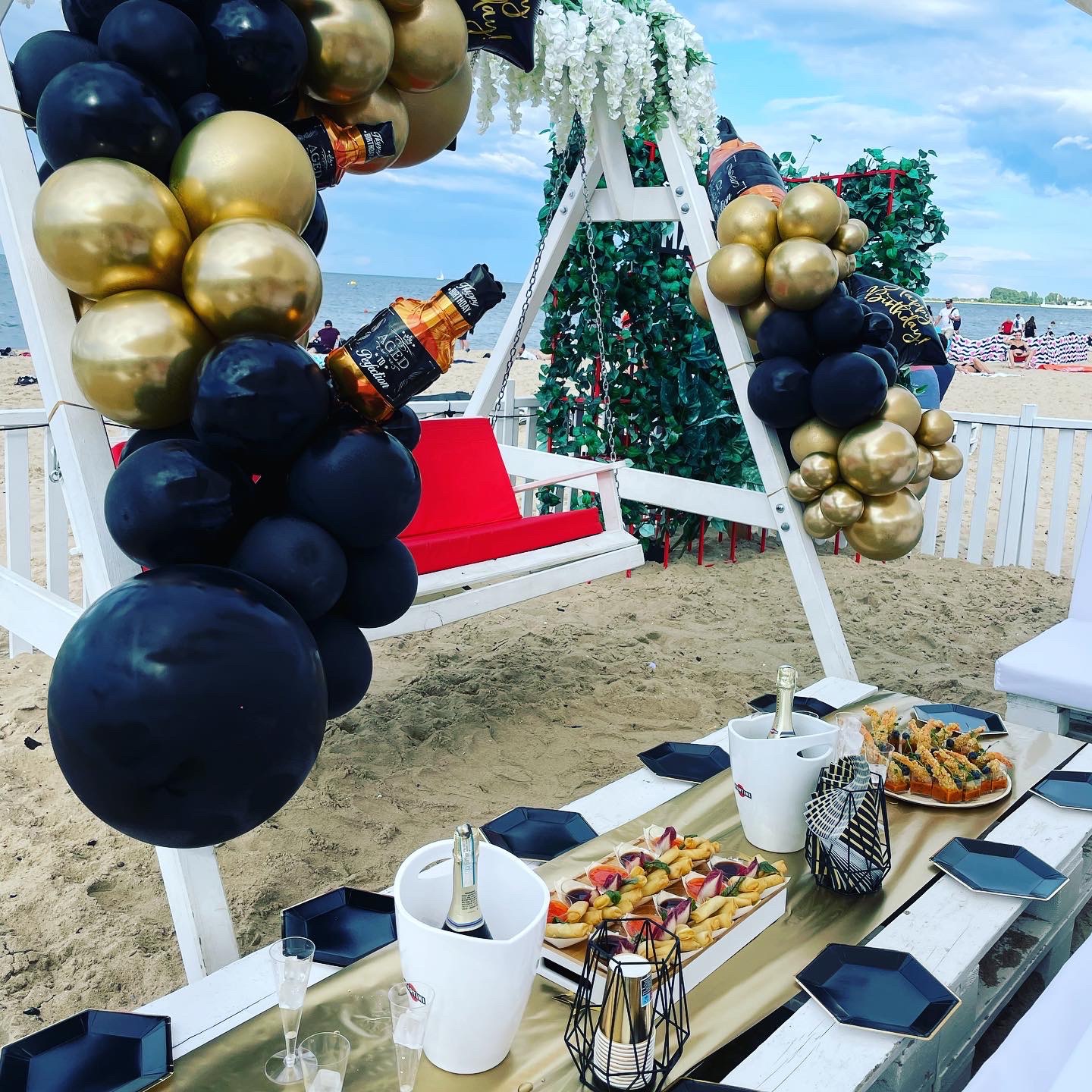 Plażowa aranżacja urodzinowa: biała huśtawka z czerwonym siedziskiem, dekoracja z czarno-złotych balonów i sztucznych kwiatów, stół z przekąskami, szampanem i czarnymi talerzami, piasek w tle.