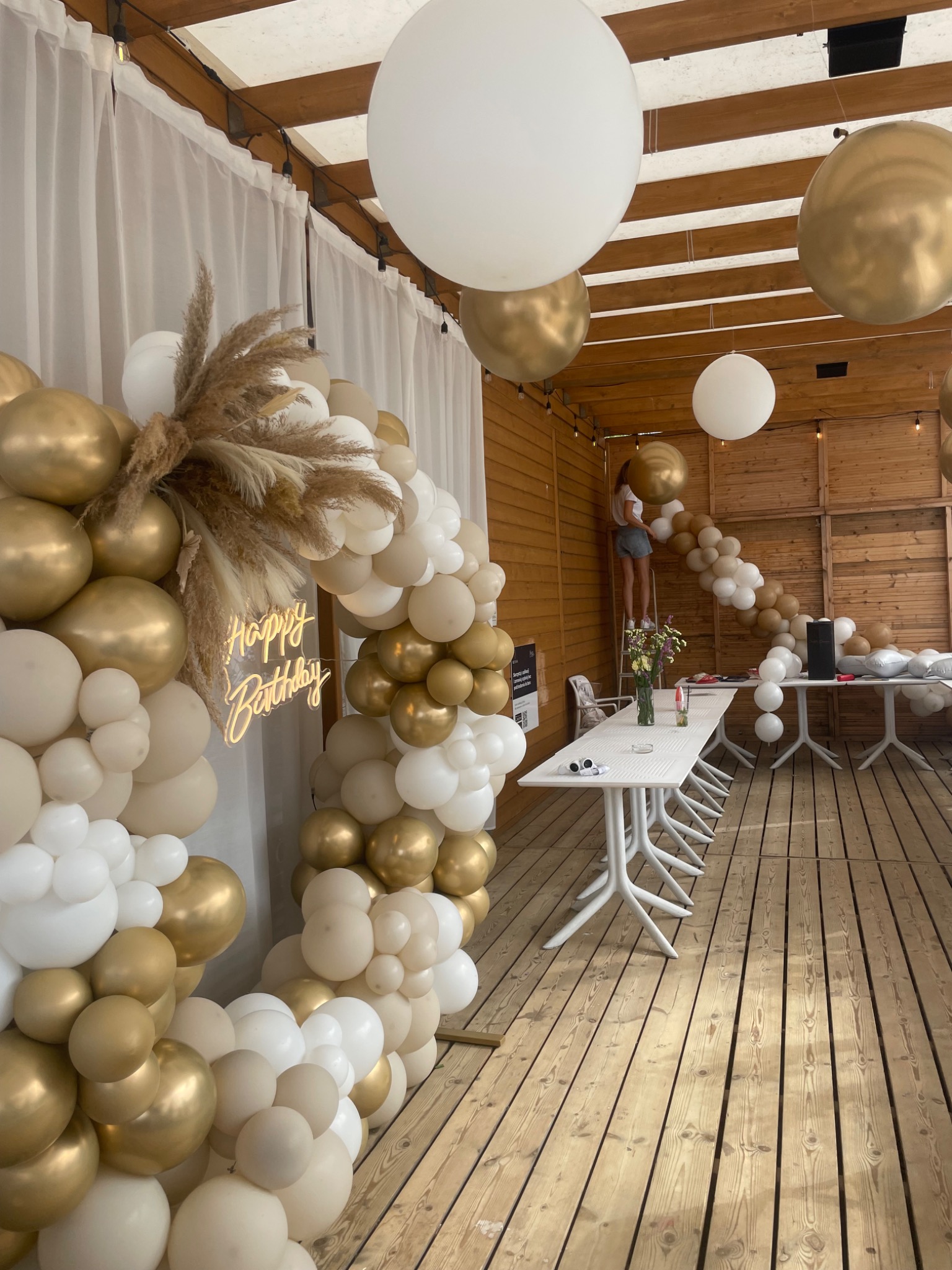 Dekoracja urodzinowa z białych, złotych i beżowych balonów, trzcin oraz neonu 'Happy Birthday' na drewnianym tarasie, osoba na drabinie mocuje girlandę balonową pod drewnianym zadaszeniem.